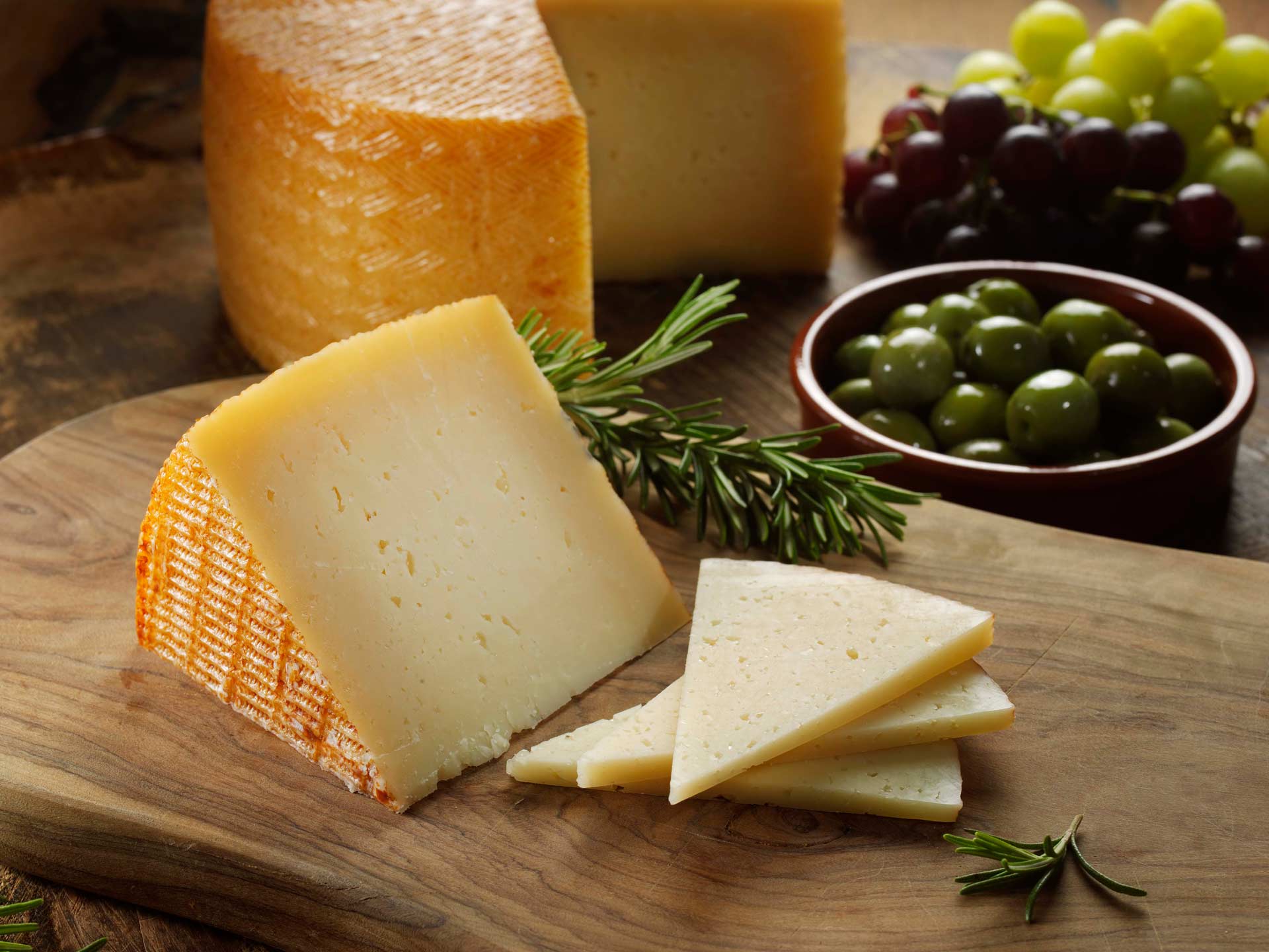 A wedge of manchego with slices cut from it. Behind them are a wheel of manchego, a bowl of olives and some grapes
