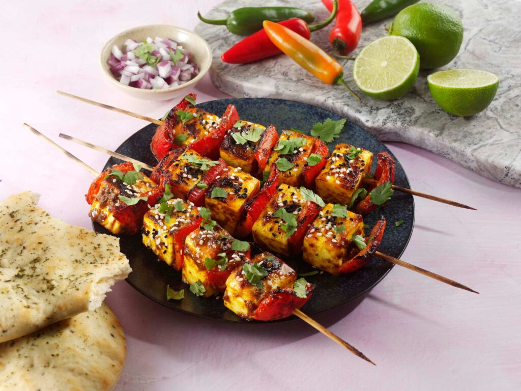 Paneer and pepper skewers with bread, red onion, limes and peppers around them