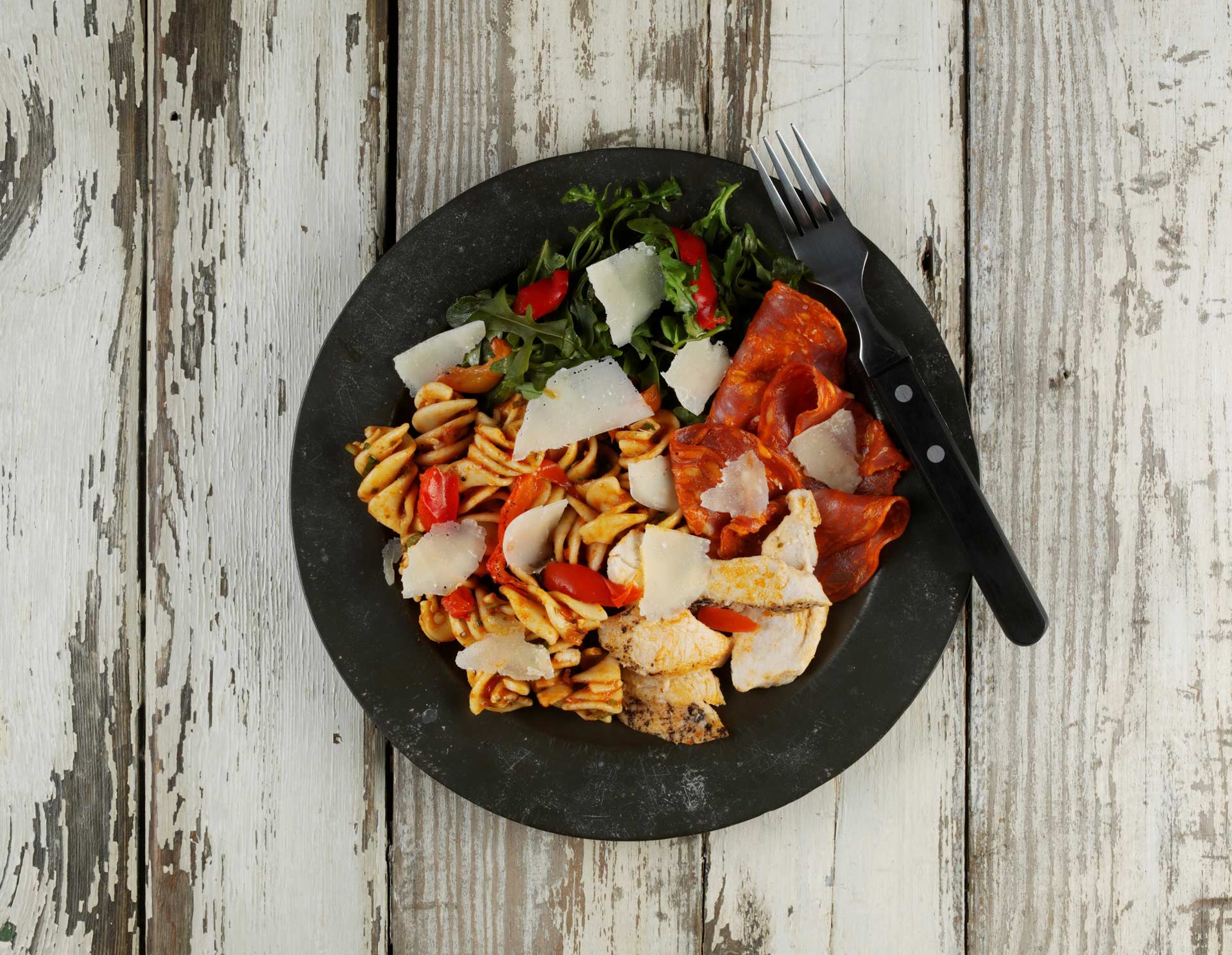 shaved grana padano pasta salad on a black plate on a distressed white painted wooden surface