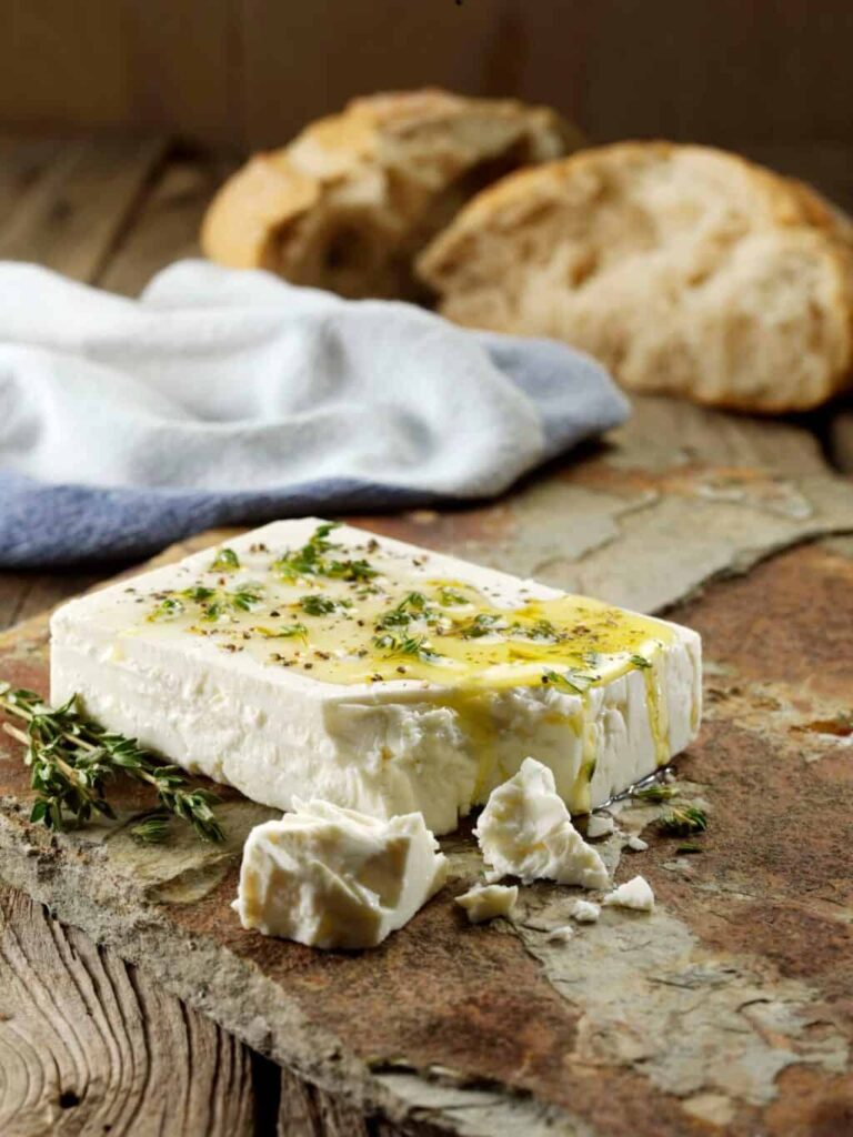 A block of feta with oil and herbs on it, and crusty bread behind