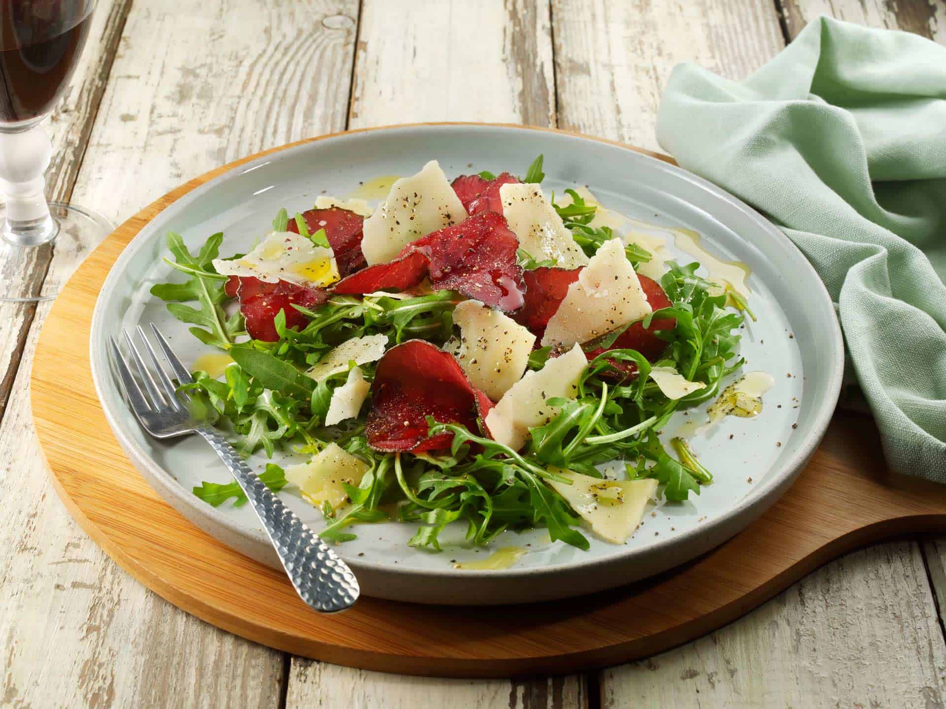 Salad with shaved Parmigiano Reggiano cheese, thinly sliced red Bresaola beef, and fresh green rocket leaves, all drizzled with olive oil and topped with black pepper.